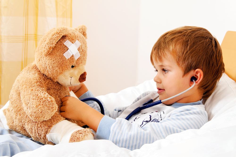 A young boy in bed using a stethoscope on his bandaged teddy bear