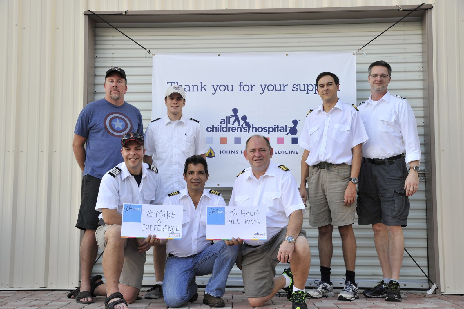 Seven Worldflight Team USA crew members, several in pilot uniforms, in front of an All Children's Hospital thank-you banner